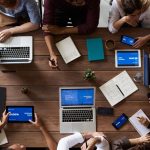 Equipe diversa reunida em torno de uma mesa, usando laptops e tablets durante uma reunião de negócios.