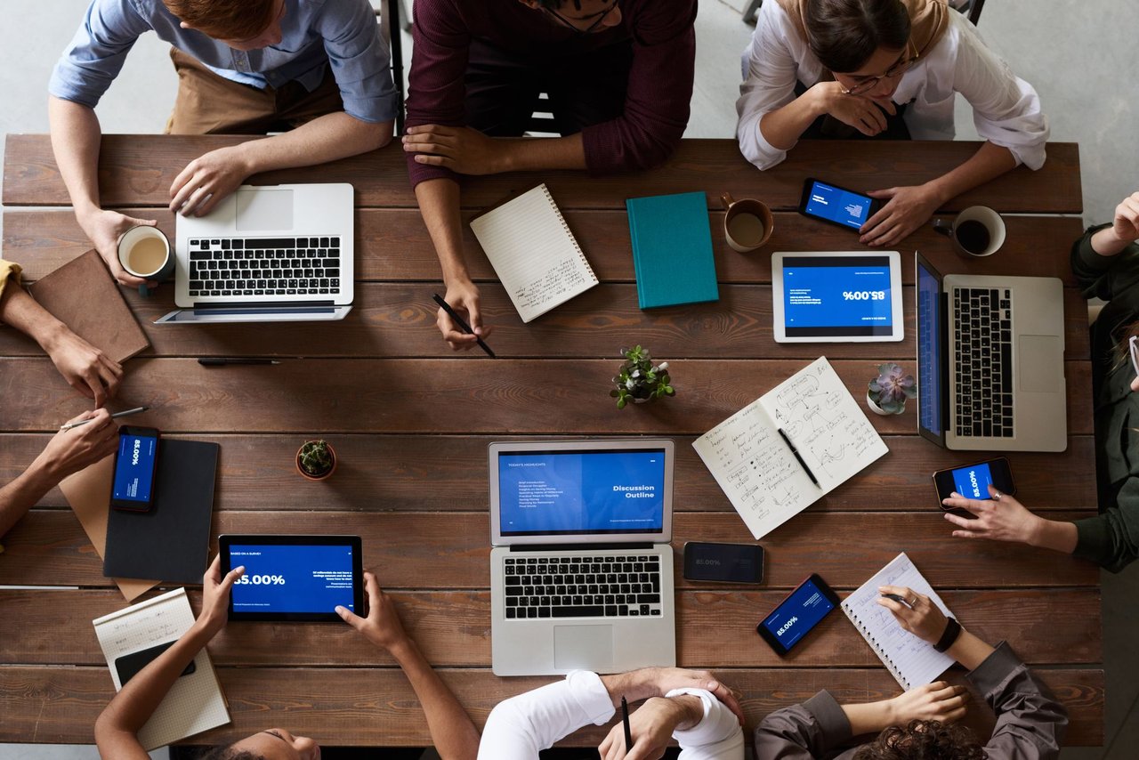 Equipe diversa reunida em torno de uma mesa, usando laptops e tablets durante uma reunião de negócios.