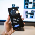 Mão segurando um smartphone com uma interface de carteira digital, diante de um monitor desfocado.