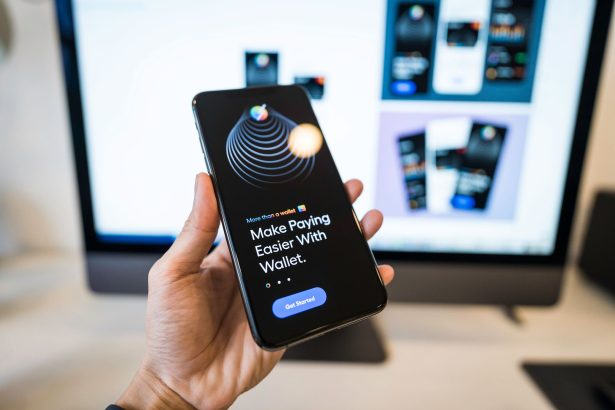 Mão segurando um smartphone com uma interface de carteira digital, diante de um monitor desfocado.